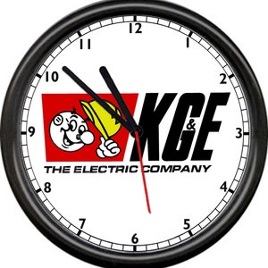 May include: Round wall clock with a black frame and a white face. The clock features the text "KGF & The Electric Company" and a cartoon character holding a yellow object. The clock hands are black, and the second hand is red.