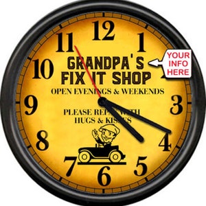 May include: A black framed clock with a yellow face. The clock face has the text "Grandpa's Fix It Shop" with the words "Open Evenings & Weekends" below. The clock face also has a cartoon image of a man driving a car. The clock has black hands and black numbers.