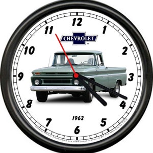 May include: A black and white wall clock with a green 1962 Chevrolet pickup truck in the center. The clock face has black numbers and a red hour hand and black minute hand. The truck has a blue Chevrolet logo on the front grill. The text "General Motors Trademarks are used under license to Off The Wall Clocks F056" is printed at the bottom of the clock.