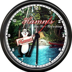 May include: A round wall clock with a black frame. The clock face features a scenic image with a bear holding a sign that says "Hamm's". The clock also has the text "The Beer Refreshing". The clock hands are black and the second hand is red.