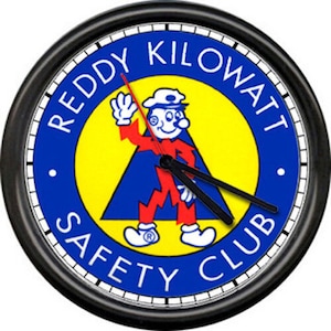 May include: A black wall clock with a white face and black hands. The clock face features a yellow and blue circle with the text "Reddy Kilowatt Safety Club" and a cartoon character with a lightning bolt.