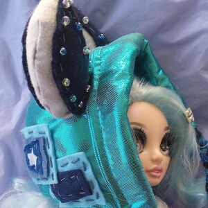 May include: A doll wearing a handmade teal and blue hood with a matching head covering. The hood is embellished with small, clear beads and patches. The doll has blue hair and is set against a blue background.