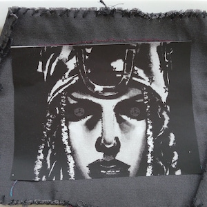 May include: A black and white print of a woman's face, featuring a detailed headpiece, is centered on a gray fabric patch. The patch has a raw, stitched edge, with visible thread. The image is a close-up, highlighting the woman's features.