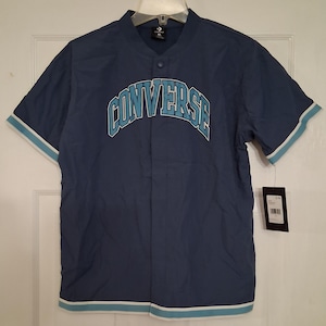 Op de afbeelding: Een marineblauw honkbalshirt met korte mouwen. Het woord "CONVERSE" staat in een boog over de borst in lichtblauw. De shirt heeft een knoopsluiting, een kraagloze halslijn en lichtblauwe afwerking op de mouwen en zoom. Een label hangt eraan.