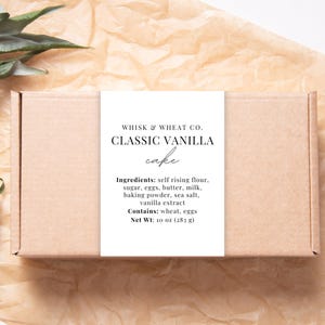 May include: A brown box with a white label that reads "WHISK & WHEAT CO. CLASSIC VANILLA cake" and lists the ingredients: self rising flour, sugar, eggs, butter, milk, baking powder, sea salt, vanilla extract. It also states "Contains: wheat, eggs" and "Net Wt: 10 oz (28 g)".