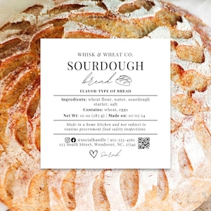 May include: A white label with black text that reads "Whisk & Wheat Co. Sourdough Bread" with a handwritten style font. The label includes the ingredients, net weight, date made, and contact information for the bakery. The label is placed on top of a loaf of sourdough bread.
