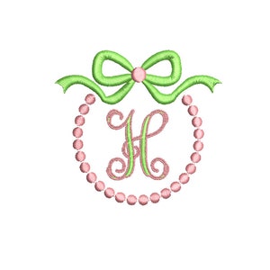 May include: A green bow with a pink center sits atop a circle of pink beads, with a green and pink letter "H" in the center.