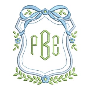 May include: Embroidered monogram design featuring the letters PBE in green, framed within a light blue shield shape. A light blue ribbon and floral accents adorn the top, with a garland of leaves and flowers at the bottom. The design is on a white background.