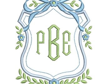 May include: Embroidered monogram design featuring the letters PBE in green, framed within a light blue shield shape. A light blue ribbon and floral accents adorn the top, with a garland of leaves and flowers at the bottom. The design is on a white background.