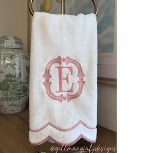 K&ouml;nnte beinhalten: Ein wei&szlig;es Handtuch mit einem rosa gestickten Monogrammbuchstaben E in einem dekorativen Rahmen. Die Unterkante des Handtuchs hat ein wellenf&ouml;rmiges Design, das mit rosa Faden eingefasst ist. Der Text @gillmangirlsdesigns ist sichtbar.