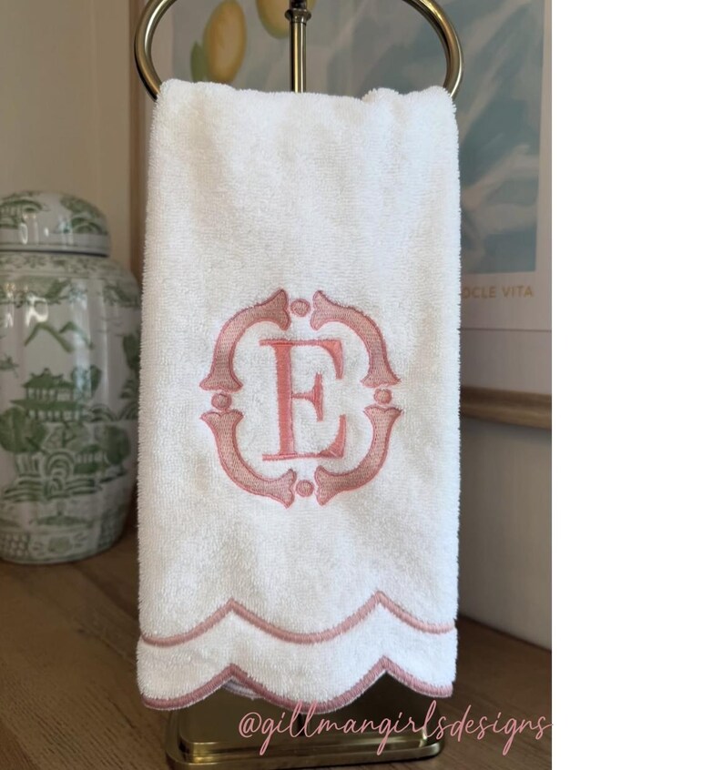 K&ouml;nnte beinhalten: Ein wei&szlig;es Handtuch mit einem rosa gestickten Monogrammbuchstaben E in einem dekorativen Rahmen. Die Unterkante des Handtuchs hat ein wellenf&ouml;rmiges Design, das mit rosa Faden eingefasst ist. Der Text @gillmangirlsdesigns ist sichtbar.