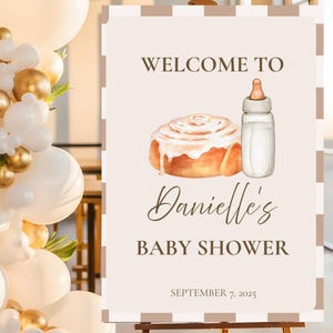 May include: A baby shower sign on an easel. The sign has a light beige background with a brown border. The sign reads "Welcome to Danielle's Baby Shower" with an illustration of a cinnamon roll and a baby bottle. The date is September 7, 2025.