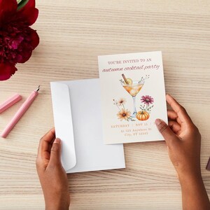 May include: An invitation for an autumn cocktail party is held in front of a white envelope. The invitation features a watercolor illustration of a cocktail, flowers, and a pumpkin. A pink pen and a red flower are also visible.