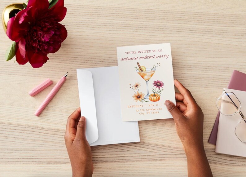 May include: An invitation for an autumn cocktail party is held in front of a white envelope. The invitation features a watercolor illustration of a cocktail, flowers, and a pumpkin. A pink pen and a red flower are also visible.