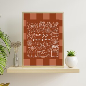 Könnte beinhalten: Ein gerahmter Druck mit einem braun-weißen karierten Hintergrund, der weiße Strichzeichnungen mit Herbstmotiven zeigt. Die Worte "cozy season" sind kursiv geschrieben. Das Bild enthält einen Kürbis, Schleifen und ein Glas Marmelade.