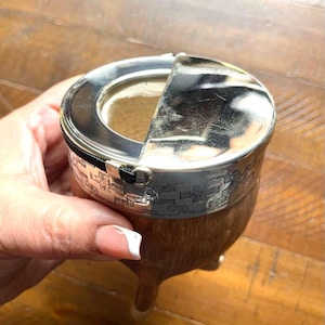 Może przedstawiać: Tykwy ze srebrną obw&oacute;dką i otwieraną pokrywką, prawdopodobnie do picia yerba mate. Tykwa ma drewnianą podstawę i tkany wz&oacute;r wok&oacute;ł srebrnej obw&oacute;dki. Pokrywka jest częściowo otwarta, odsłaniając wnętrze.