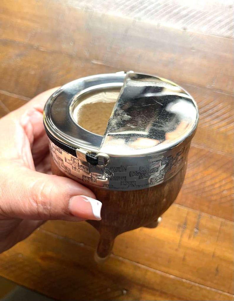 Może przedstawiać: Tykwy ze srebrną obw&oacute;dką i otwieraną pokrywką, prawdopodobnie do picia yerba mate. Tykwa ma drewnianą podstawę i tkany wz&oacute;r wok&oacute;ł srebrnej obw&oacute;dki. Pokrywka jest częściowo otwarta, odsłaniając wnętrze.