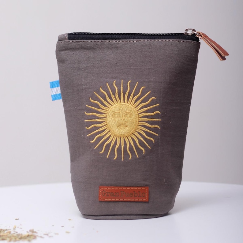 Op de afbeelding: Een grijze stoffen etui met een ritssluiting en een leren treklipje. De etui heeft een geborduurd gouden zonontwerp en een kleine Argentijnse vlag. Een bruin rechthoekig label met tekst is aan de onderkant genaaid.