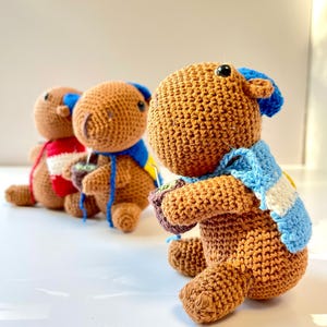 Op de afbeelding: Drie handgemaakte gehaakte capibara's op een rij, elk met unieke accessoires. De voorste capibara is bruin met een blauw-witte sjaal en een kleine pot. De andere hebben verschillend gekleurde sjaals.
