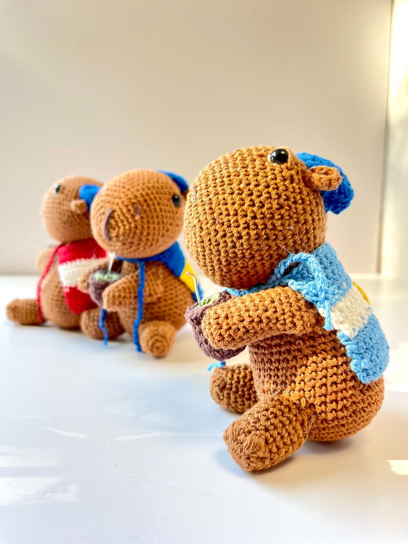 Op de afbeelding: Drie handgemaakte gehaakte capibara's op een rij, elk met unieke accessoires. De voorste capibara is bruin met een blauw-witte sjaal en een kleine pot. De andere hebben verschillend gekleurde sjaals.