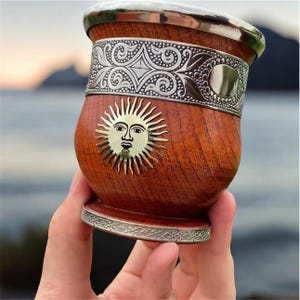 Könnte beinhalten: Ein Yerba-Mate-Gefäß aus Holz mit einem dekorativen Metallband und -rand. Das Gefäß hat ein goldenes Sonnenemblem und wird in einer Hand gehalten. Der Hintergrund ist verschwommen, was auf eine Umgebung im Freien hindeutet.