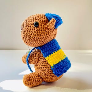 Op de afbeelding: Een gehaakte bruine berenpop met een blauw-gele gestreepte sjaal en een blauwe hoed. De handgemaakte beer heeft zwarte ogen en zit. De pop is gemaakt van garen en heeft een getextureerd uiterlijk.