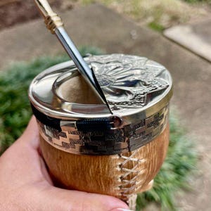 Może przedstawiać: Ręcznie wykonana tykwa do yerba mate ze srebrną obw&oacute;dką i pokrywką, z wytłoczonym wzorem. Tykwa ma naturalny brązowy wygląd zewnętrzny z ozdobnymi szwami i czarnym paskiem. Włożona jest srebrna słomka bombilla.