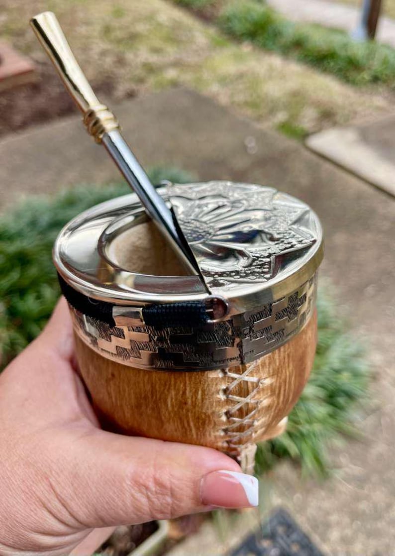 Może przedstawiać: Ręcznie wykonana tykwa do yerba mate ze srebrną obw&oacute;dką i pokrywką, z wytłoczonym wzorem. Tykwa ma naturalny brązowy wygląd zewnętrzny z ozdobnymi szwami i czarnym paskiem. Włożona jest srebrna słomka bombilla.