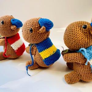 Op de afbeelding: Drie handgemaakte gehaakte berenpoppen. Elke beer is bruin met een blauwe hoed en een sjaal in verschillende kleuren: rood en wit, blauw en geel en lichtblauw en wit. De beren zitten.