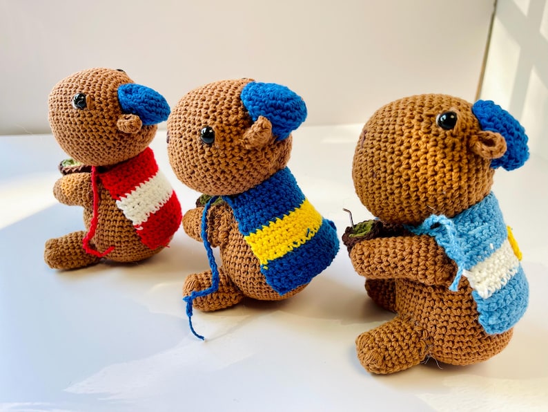 Op de afbeelding: Drie handgemaakte gehaakte berenpoppen. Elke beer is bruin met een blauwe hoed en een sjaal in verschillende kleuren: rood en wit, blauw en geel en lichtblauw en wit. De beren zitten.