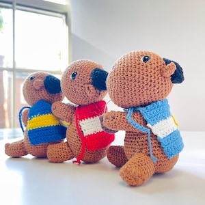 Op de afbeelding: Drie handgemaakte gehaakte hondenspeeltjes op een rij. Elke hond is bruin met zwarte oren en neus. Ze dragen kleurrijke sjaals: blauw met gele strepen, rood en wit, en blauw met witte strepen. De speeltjes staan op een wit oppervlak.