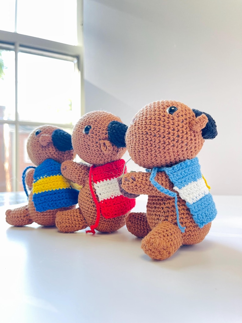 Op de afbeelding: Drie handgemaakte gehaakte hondenspeeltjes op een rij. Elke hond is bruin met zwarte oren en neus. Ze dragen kleurrijke sjaals: blauw met gele strepen, rood en wit, en blauw met witte strepen. De speeltjes staan op een wit oppervlak.