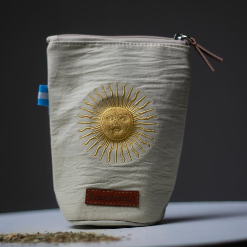 Op de afbeelding: Een beige canvas etui met een ritssluiting en een bruine leren trekker. De etui heeft een goud geborduurd zonontwerp en een klein blauw-wit label. Een bruin leren label aan de onderkant leest "Gran Pueblo."