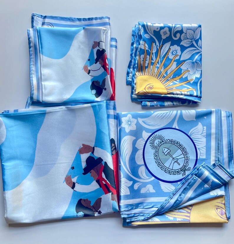 May include: Several folded silk scarves in shades of light blue and white are displayed. The scarves feature various designs, including illustrations of people, floral patterns, and a sun with a face. The edges of the scarves have a striped border.