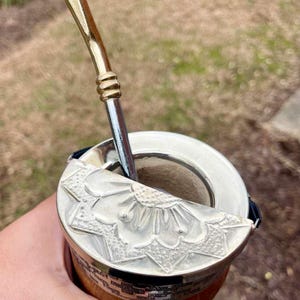 Może przedstawiać: Srebrny kubek do yerba mate z ozdobną pokrywką i metalową słomką. Kubek ma drewnianą podstawę i srebrną obręcz. Słomka ma złotą g&oacute;rę.