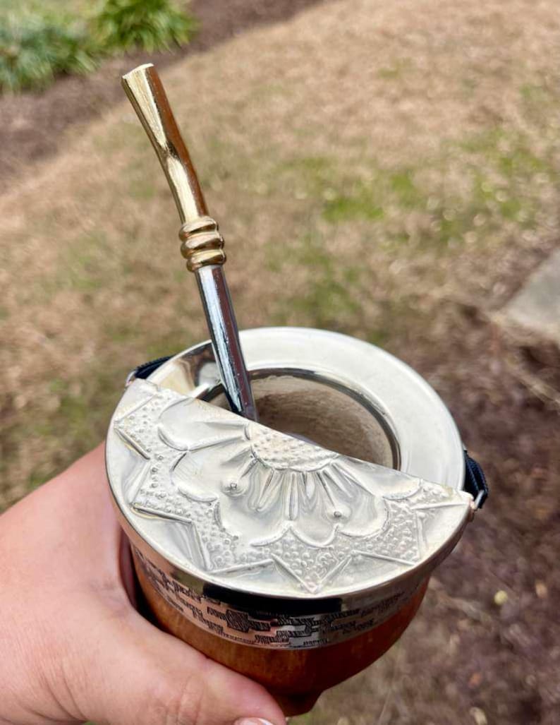 Może przedstawiać: Srebrny kubek do yerba mate z ozdobną pokrywką i metalową słomką. Kubek ma drewnianą podstawę i srebrną obręcz. Słomka ma złotą g&oacute;rę.