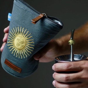 Op de afbeelding: Een grijze ge&iuml;soleerde yerba mate gourd houder met een gouden geborduurd zonontwerp. De houder heeft een ritssluiting en een bruine leren trekker. Een metalen yerba mate beker en rietje zijn ook zichtbaar.