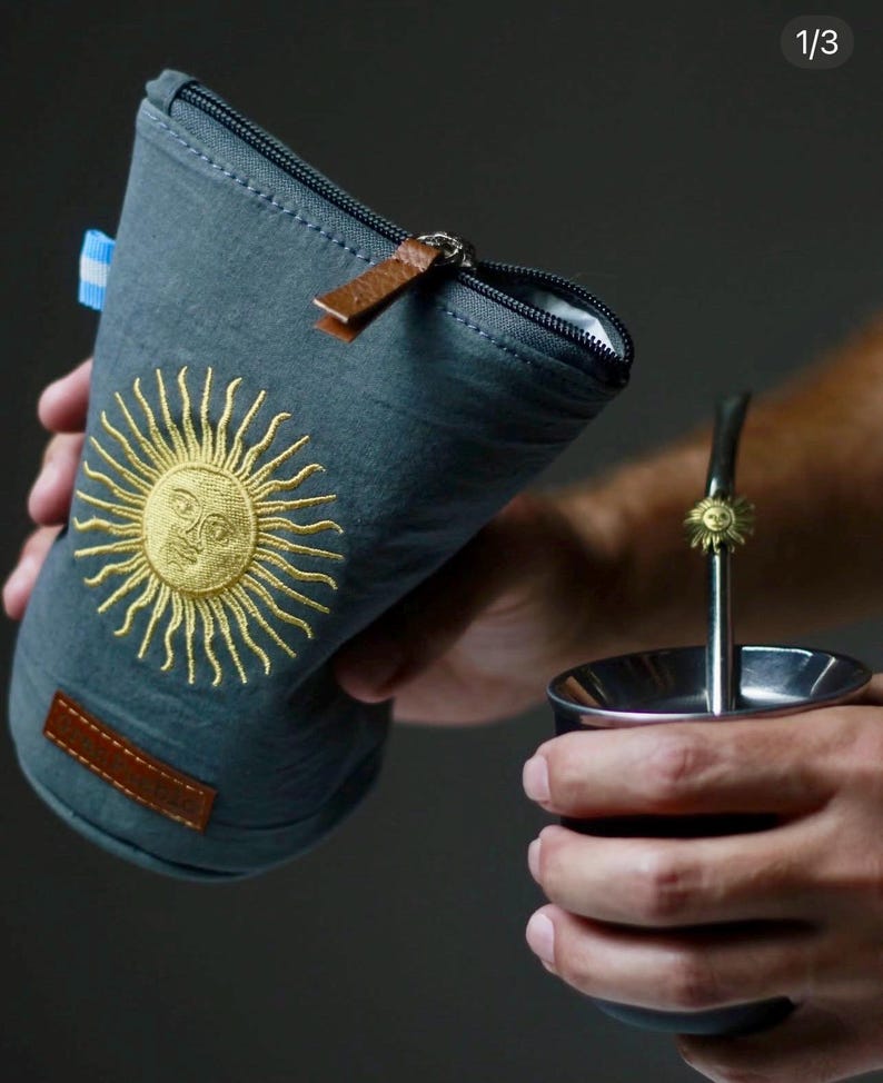 Op de afbeelding: Een grijze ge&iuml;soleerde yerba mate gourd houder met een gouden geborduurd zonontwerp. De houder heeft een ritssluiting en een bruine leren trekker. Een metalen yerba mate beker en rietje zijn ook zichtbaar.