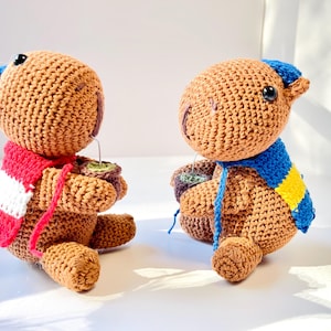 Op de afbeelding: Twee handgemaakte gehaakte teddyberen, &eacute;&eacute;n met een rood-witte sjaal, de andere met een blauw-gele sjaal. Elke beer houdt een klein kopje met een groene drank vast. De beren zijn bruin met blauwe accenten.