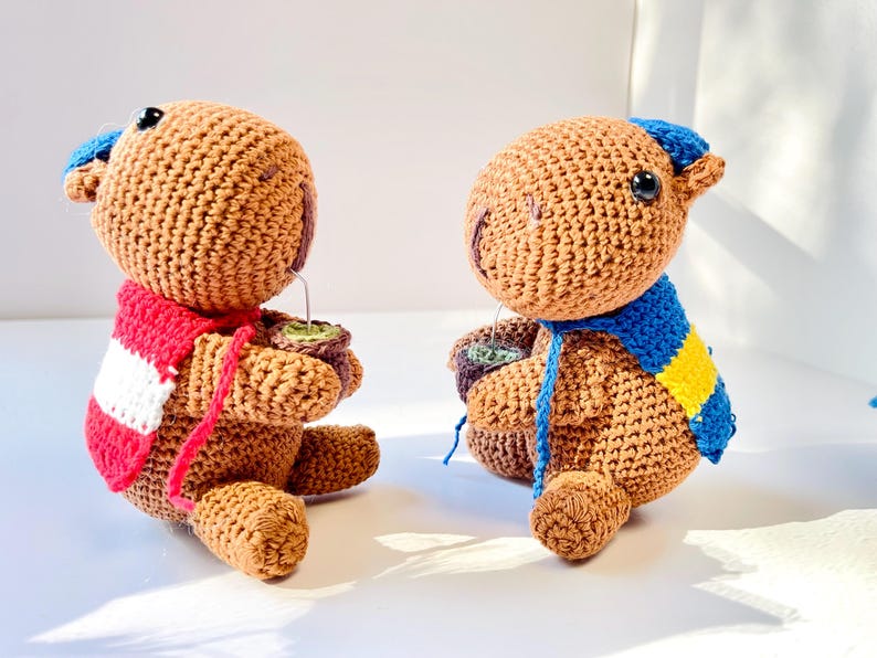 Op de afbeelding: Twee handgemaakte gehaakte teddyberen, &eacute;&eacute;n met een rood-witte sjaal, de andere met een blauw-gele sjaal. Elke beer houdt een klein kopje met een groene drank vast. De beren zijn bruin met blauwe accenten.