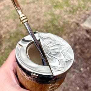 Może przedstawiać: Tykwy do yerba mate ze srebrną obręczą i pokrywką, z wytłoczonym kwiatowym wzorem. Srebrna i złota słomka bombilla jest włożona do tykwy. Tykwa ma naturalne drewniane wykończenie i czarny zamek błyskawiczny.