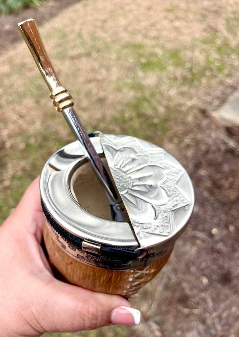 Może przedstawiać: Tykwy do yerba mate ze srebrną obręczą i pokrywką, z wytłoczonym kwiatowym wzorem. Srebrna i złota słomka bombilla jest włożona do tykwy. Tykwa ma naturalne drewniane wykończenie i czarny zamek błyskawiczny.