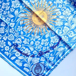 May include: A square, light blue scarf with a floral pattern and a gold sun design. The scarf has white floral accents and a border. Text in blue script reads "Coronados de Gloria Vivamos".