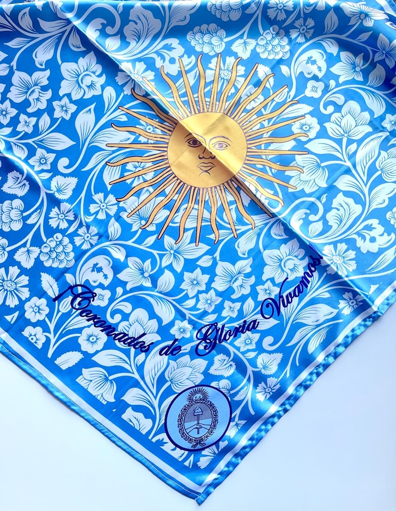 May include: A square, light blue scarf with a floral pattern and a gold sun design. The scarf has white floral accents and a border. Text in blue script reads "Coronados de Gloria Vivamos".