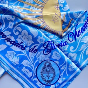 May include: A blue and white patterned scarf with the text "Vivamos de Gloria" and a sun graphic. The scarf also features a circular emblem with a wheat and tools design.