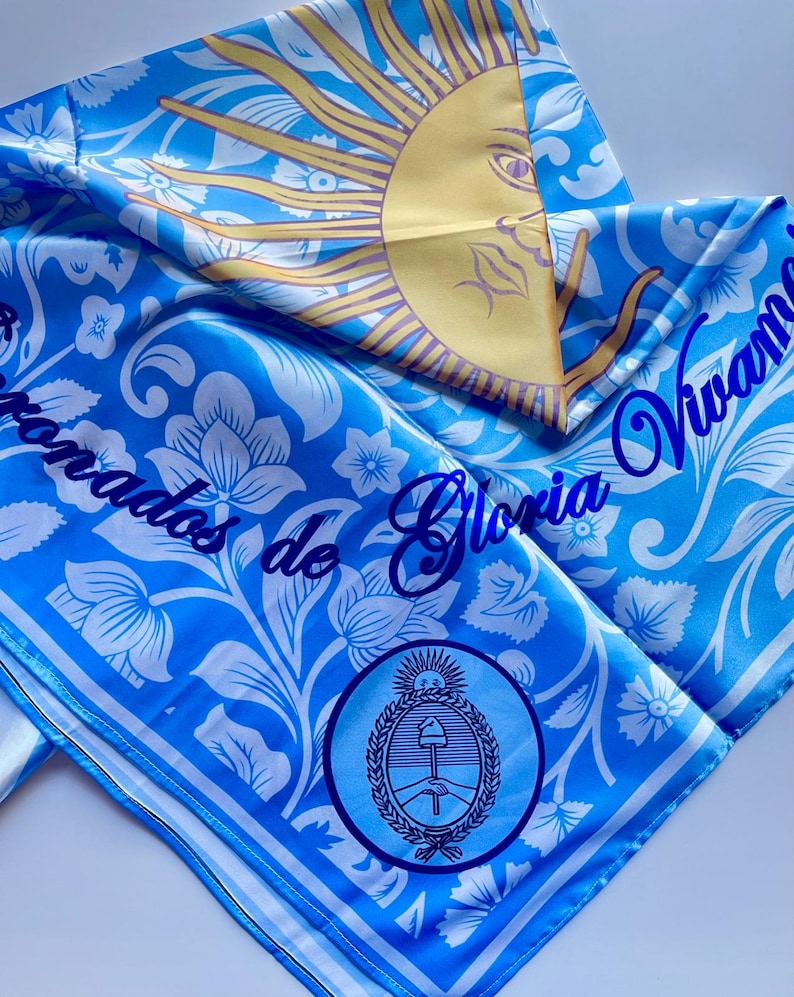 May include: A blue and white patterned scarf with the text "Vivamos de Gloria" and a sun graphic. The scarf also features a circular emblem with a wheat and tools design.