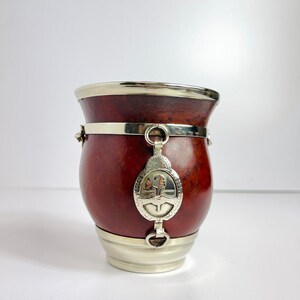 May include: A traditional yerba mate gourd with a dark red exterior and silver-colored metal accents. The gourd features a decorative metal emblem and a silver band around the middle. The base and rim are also silver.