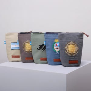 Op de afbeelding: Een rij van zes cilindrische stoffen zakjes in verschillende kleuren, elk met geborduurde ontwerpen. De ontwerpen omvatten vlaggen, zonnen en een kaart. Elk zakje heeft een ritssluiting en een klein leren label.