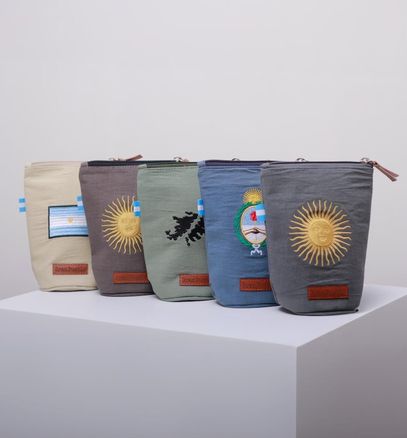 Op de afbeelding: Een rij van zes cilindrische stoffen zakjes in verschillende kleuren, elk met geborduurde ontwerpen. De ontwerpen omvatten vlaggen, zonnen en een kaart. Elk zakje heeft een ritssluiting en een klein leren label.