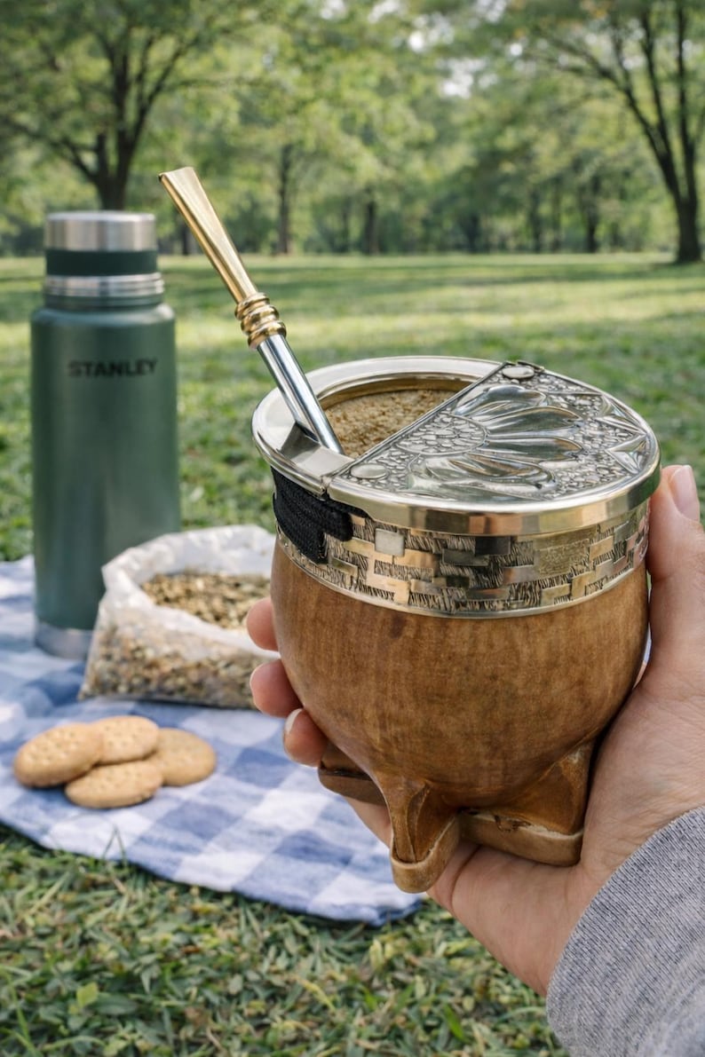 Może przedstawiać: Drewniana tykwa do yerba mate ze srebrną obręczą i pokrywką, trzymana w dłoni. Metalowa słomka jest włożona do tykwy. Zielony termos Stanley, worek yerba mate i ciastka znajdują się w tle.
